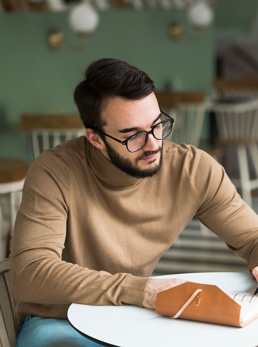 Man with glasses writing in a notebook at a café table.