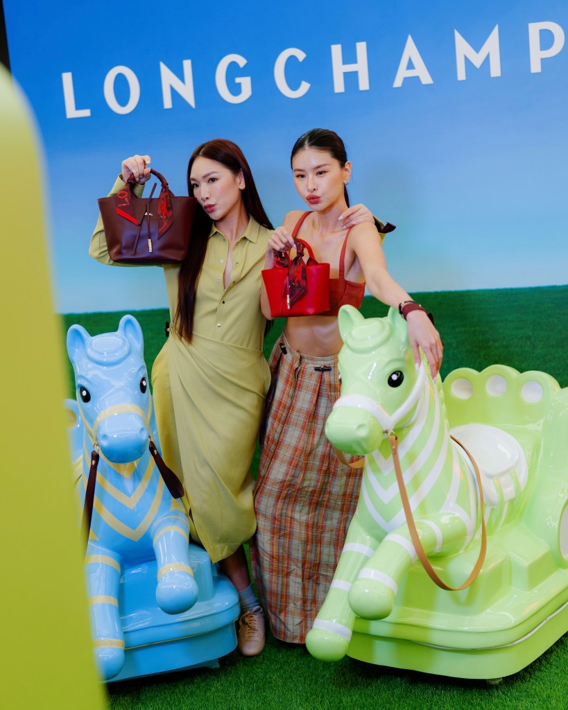 Two women posing with handbags in front of a colorful backdrop at a Longchamp event, standing next to playful horse ride-on toys.