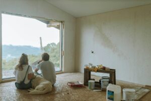 Couple seated on an unfinished floor with painting tools and a dog nearby, capturing a candid moment during a home renovation project with natural light pouring in from open French doors