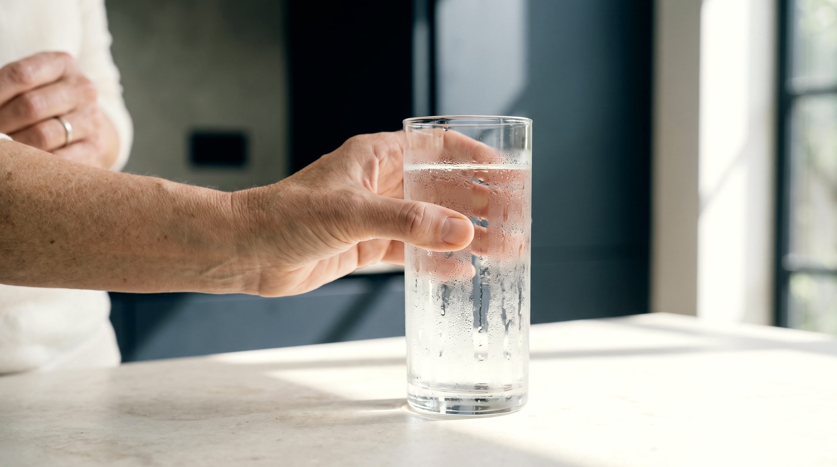 Hand hält ein beschlagenes Glas Wasser auf einer hellen Steinplatte
