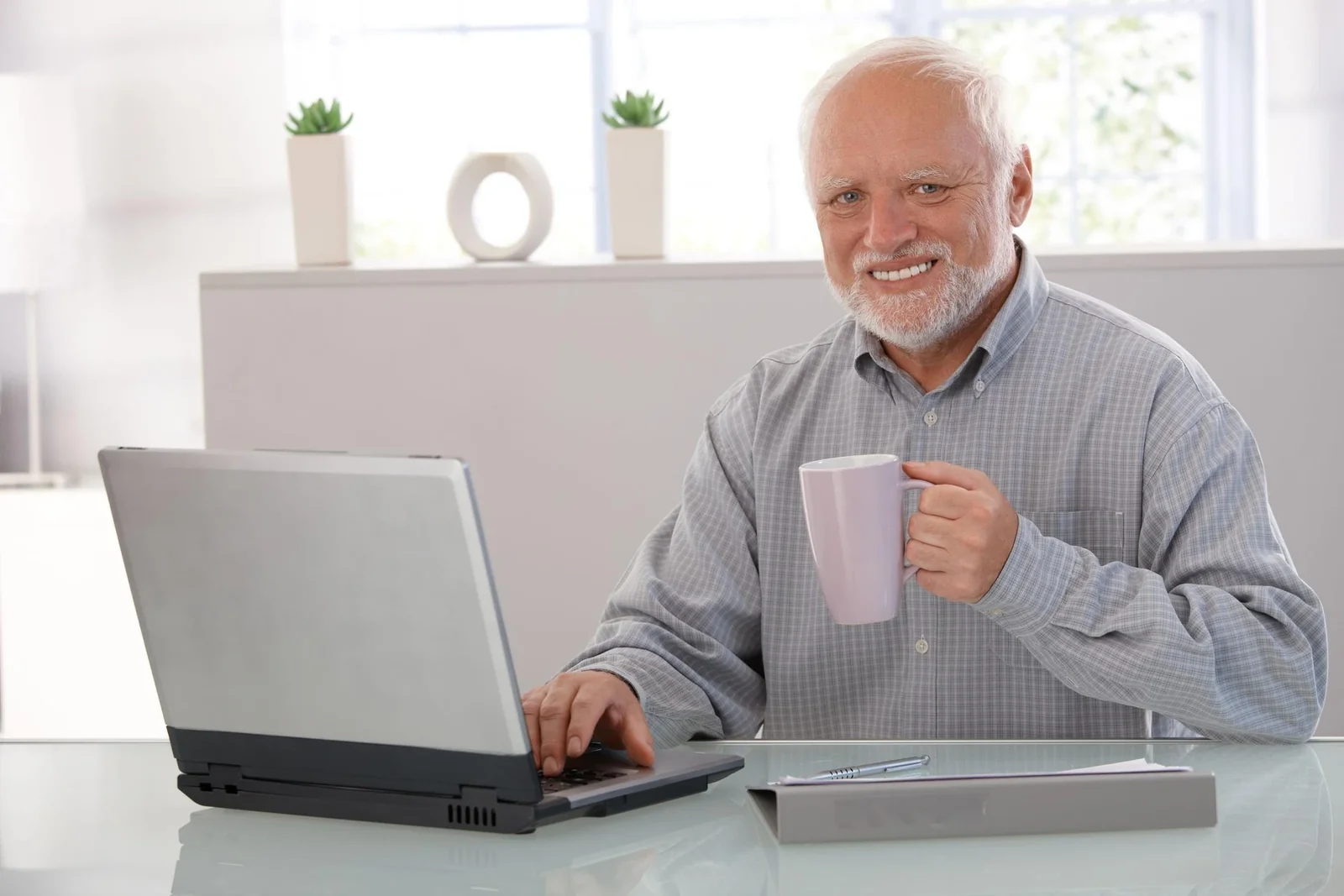 Smiling older man at desk with laptop and coffee mug, bright office setting with plants and decorative items.