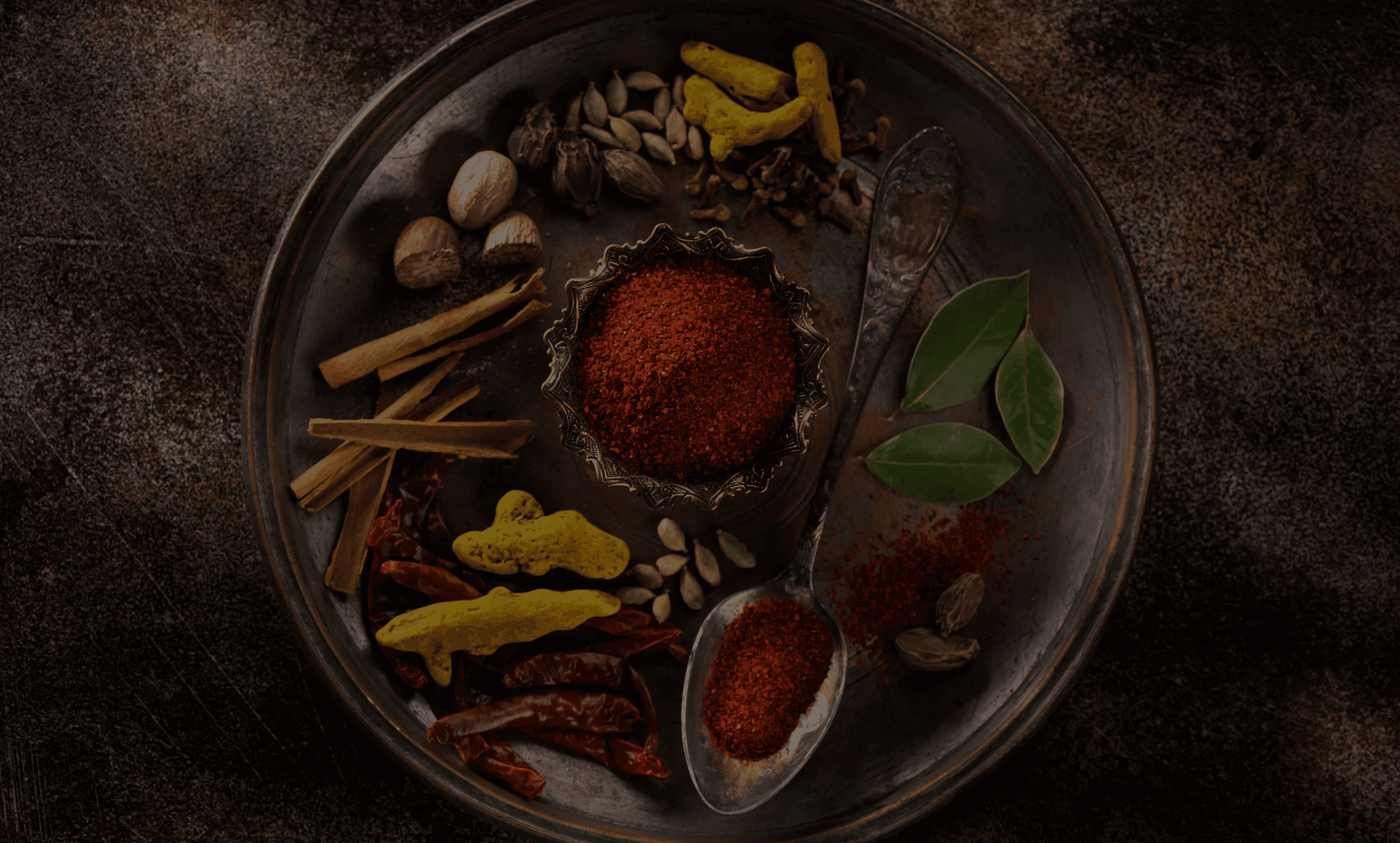 An antique metal plate displaying an array of traditional spices, including cinnamon sticks, dried red chilies, nutmeg, cardamom pods, bay leaves, cloves, black cardamom, dried turmeric, and a bowl of vibrant red chili powder with a vintage spoon beside it.