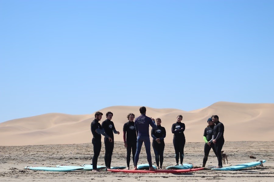 Surf instructor talking to a group in wetsuits on a beach.