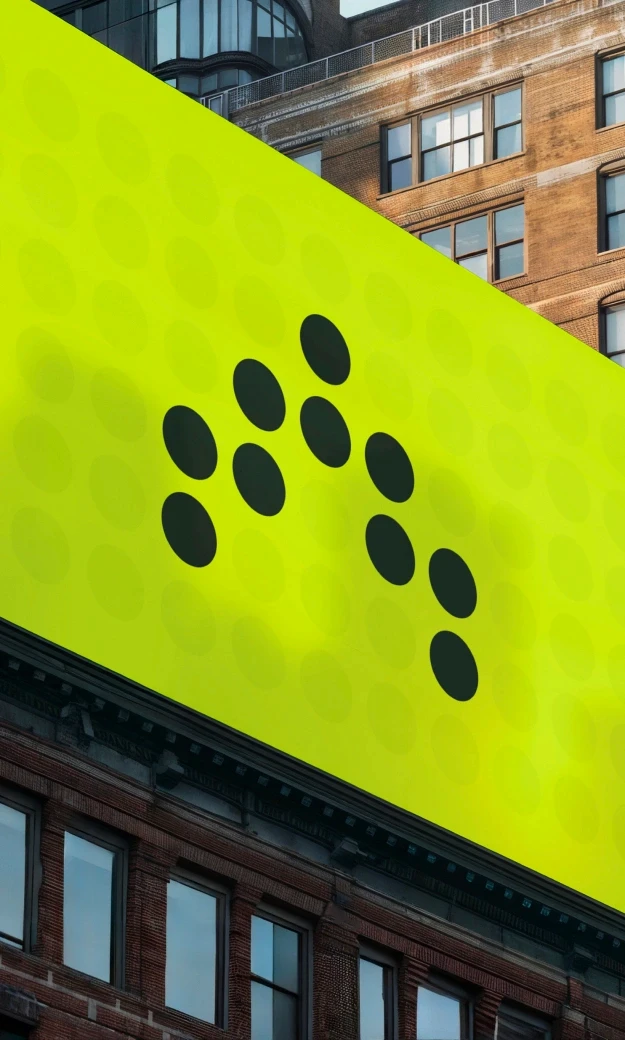 A large, bright green billboard with a minimalist design featuring black dots arranged in a pattern, displayed on the side of a tall urban building. The billboard stands out against the surrounding brick and concrete structures, with windows and architectural details visible.