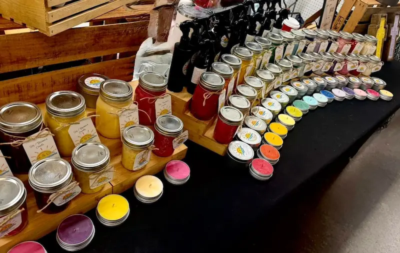 A colorful display of artisanal candles in glass jars and tins is arranged on a wooden market table in Bradenton, FL, featuring a wide array of scents and hues, including yellow, red, and purple.
