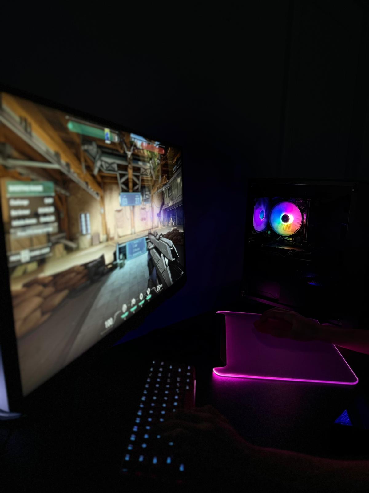 A gamer immersed in a gaming session, with hands on a backlit keyboard and mouse, while the game is being displayed on a monitor. The gaming setup includes RGB lighting inside the PC case, enhancing the immersive experience in a dark room.