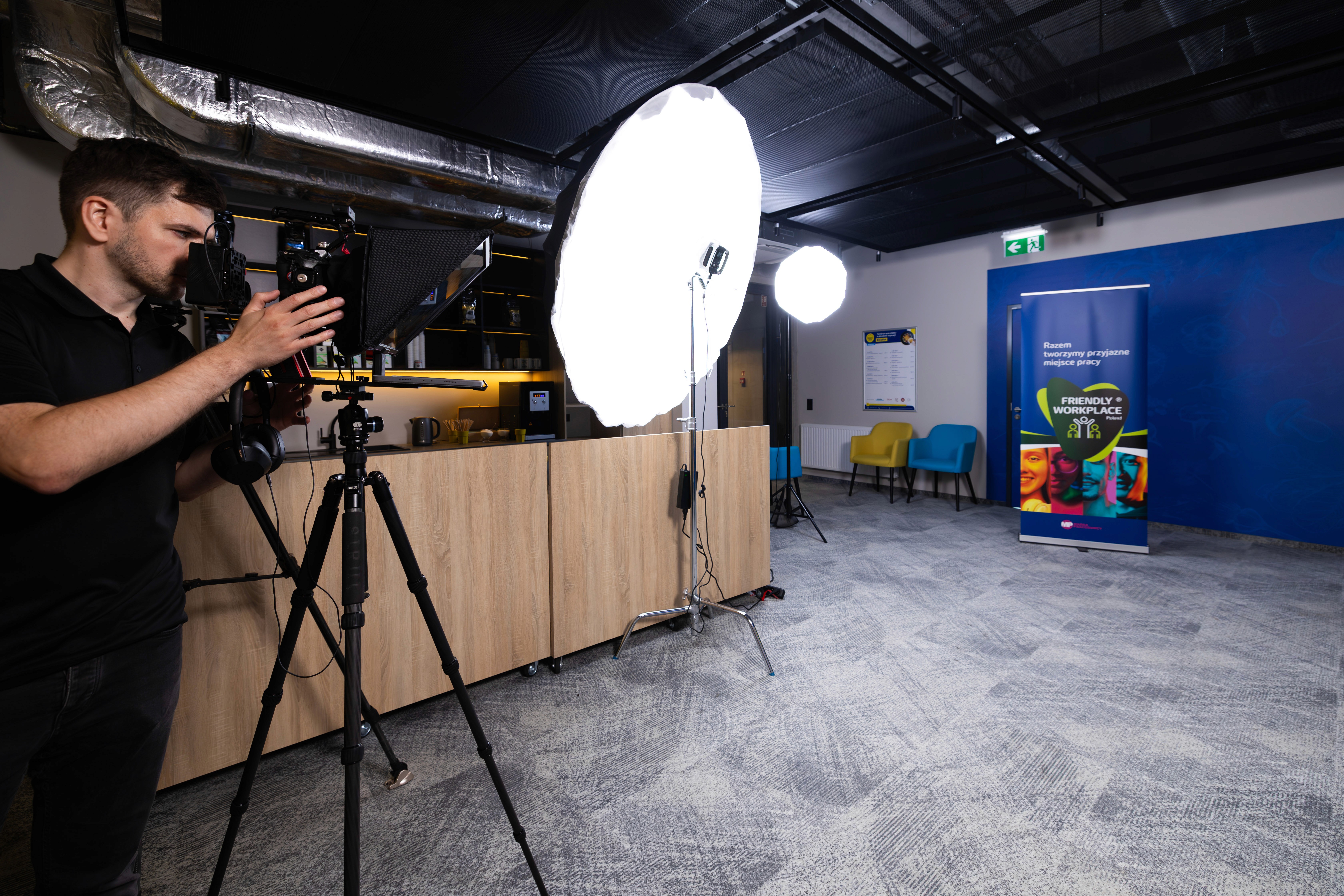 Operator kamery ustawiający profesjonalny sprzęt filmowy w biurze klienta. Widoczna wysokiej jakości kamera na statywie oraz profesjonalne oświetlenie - dwa duże softboxy na statywach. Przestrzeń biurowa zaaranżowana do nagrań z teleprompterem i innym specjalistycznym wyposażeniem, zapewniającym produkcję materiałów wideo najwyższej jakości.