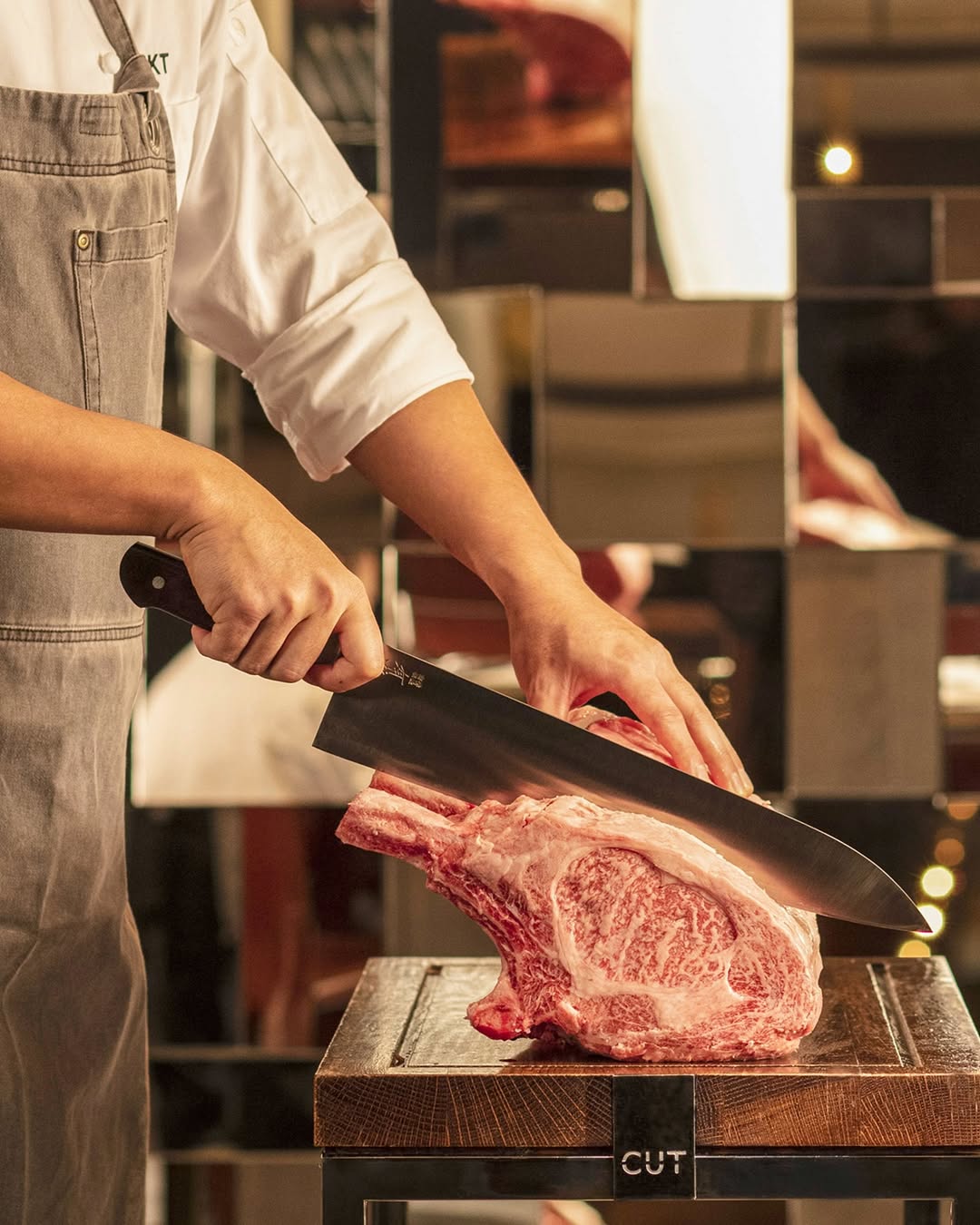 A chef wearing an apron slices a large piece of marbled meat with a sharp knife on a wooden cutting board.