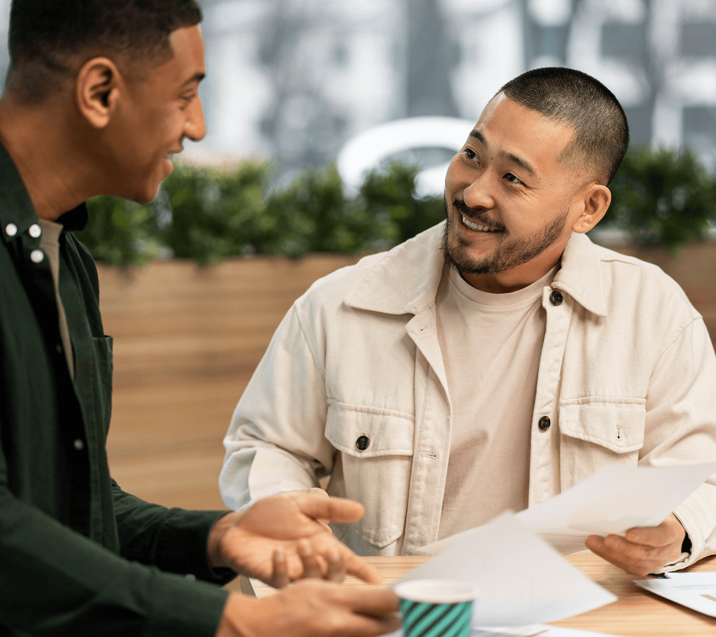 Two men in a friendly discussion over papers at a cafe table. One man smiles warmly while listening to the other speak.