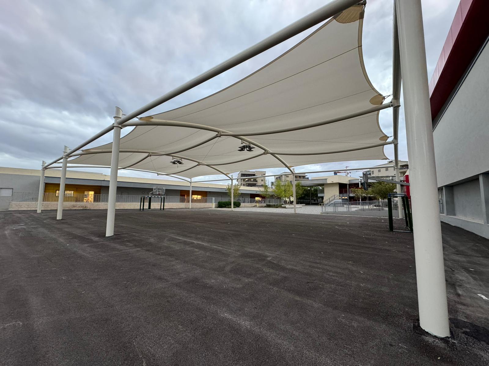 Large beige shade sails on metal frame over paved area in front of modern buildings under cloudy sky.