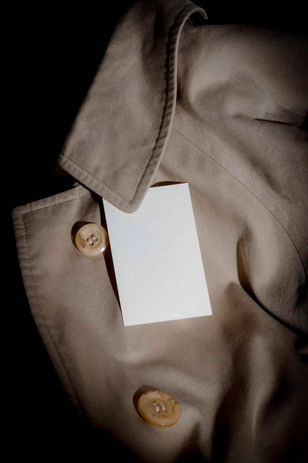 Blank white card resting on a beige trench coat with a close-up view of the fabric texture and buttons. The lighting creates dramatic shadows, emphasizing the neutral tones.