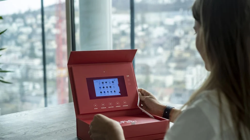 Person using a red innovation kit box with a built-in digital screen at a desk in front of a window overlooking a cityscape, symbolizing creativity and innovation tools.
