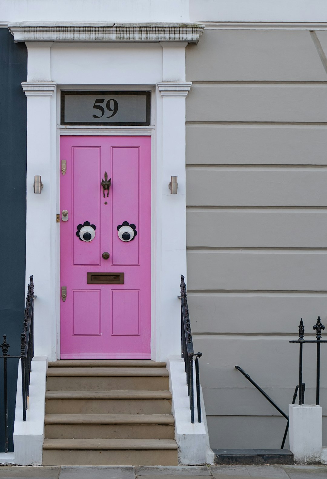 A vibrant pink front door with whimsical googly eyes sits at the top of a short flight of stairs, framed by a classic white façade and numbered "59," blending playful and traditional architectural elements in a charming residential area.