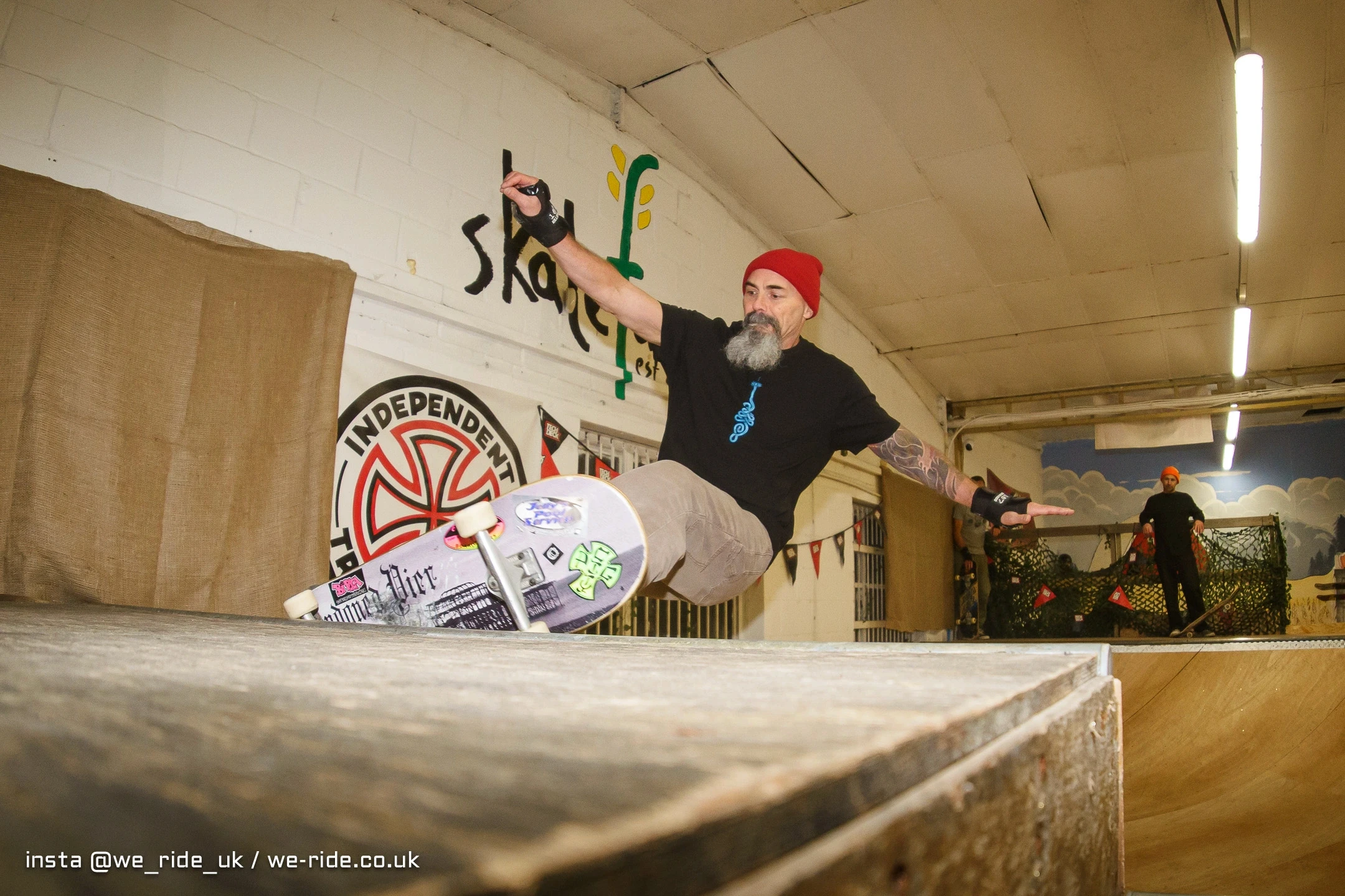 Skateboarder performs a trick in a skatepark, mid-air above a ramp, with a focused expression.