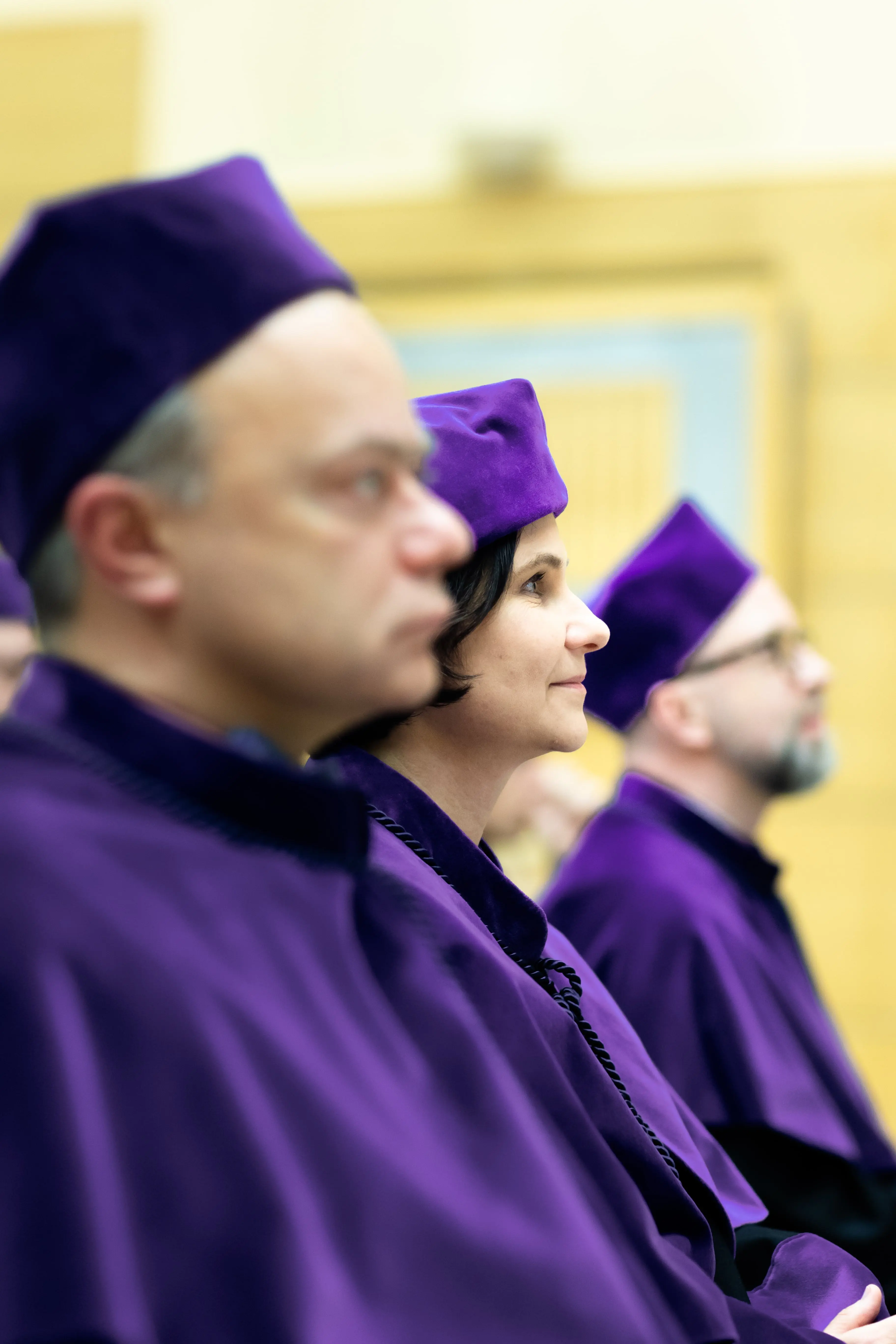 Dwójka mężczyzn i kobieta, ubrani w rektorskie togi uczelniane. SIedzą na ławie spoglądając z powagą przed siebie.