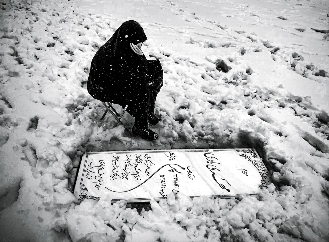 A woman in a black chador sitting on a folding chair beside a snow-covered grave with Persian script on the headstone, black and white
