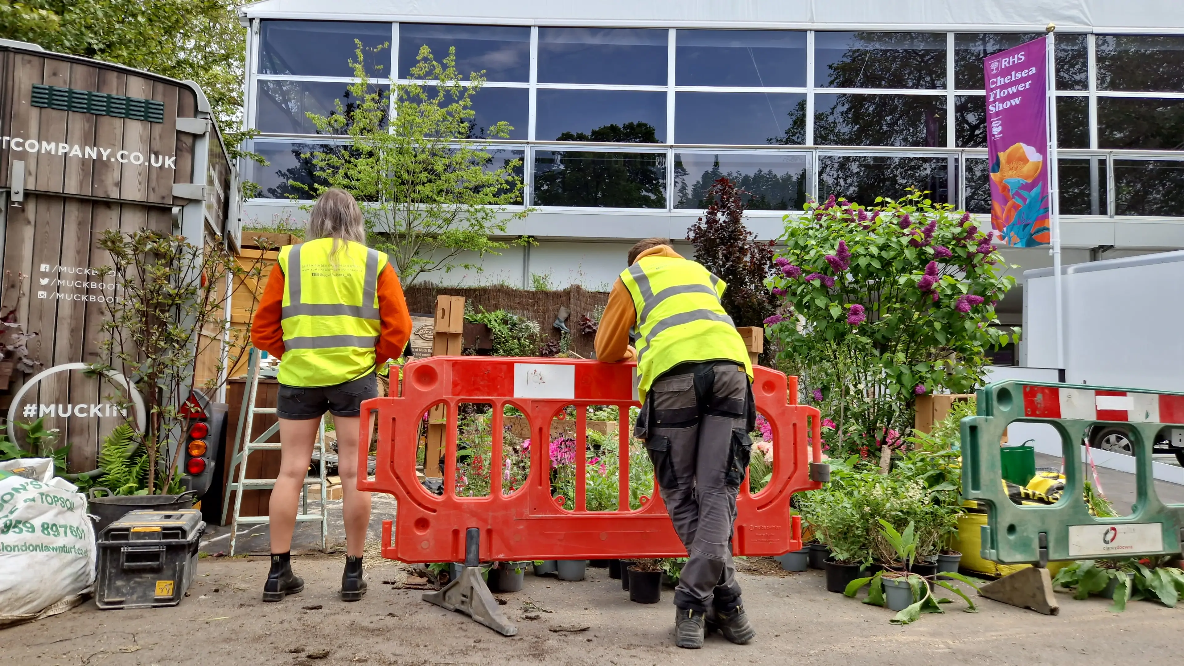 Two workers in yellow vests move a barrier in front of a building surrounded by greenery.