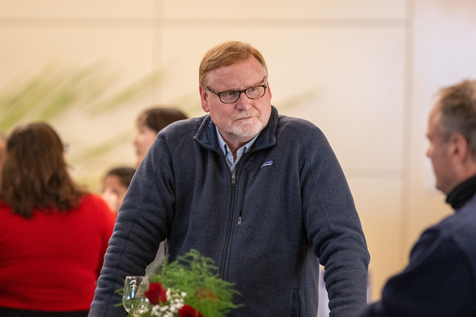 A man with glasses and a beard stands at a table, interacting with others in a casual setting.