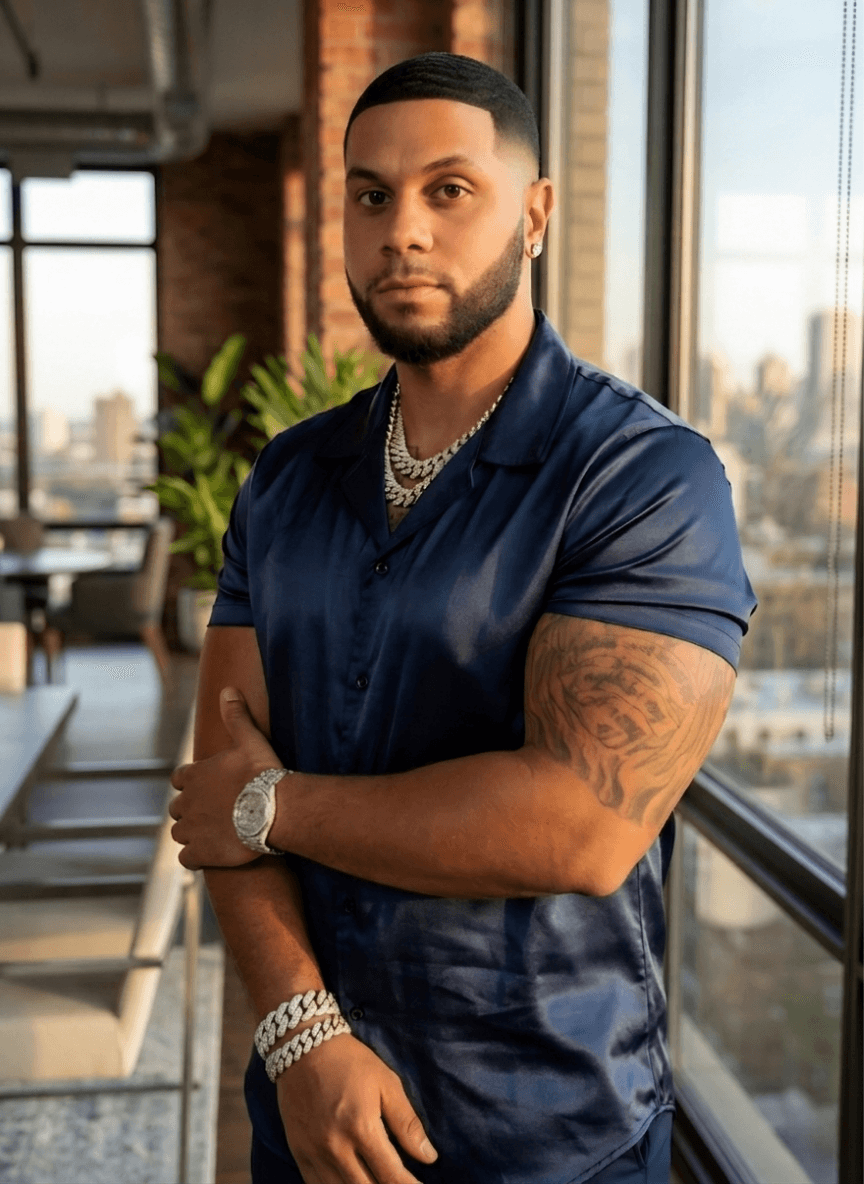 A man in a navy silk shirt stands confidently in a modern room with large windows. He has tattoos, and wears a watch and chain, conveying a stylish and relaxed vibe.