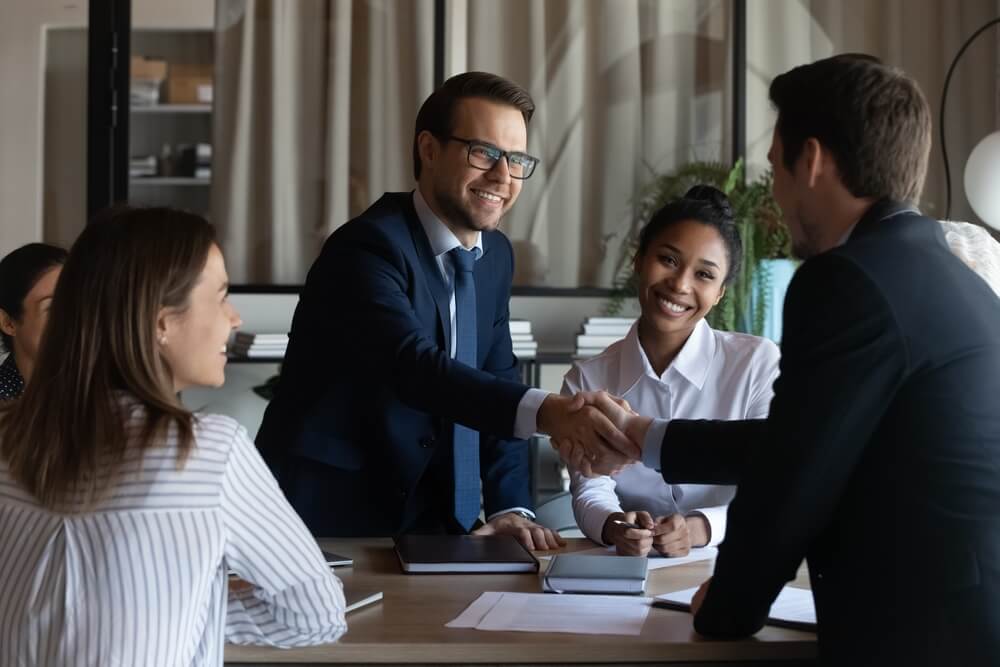 Two men shaking hands with women smiling around them in office