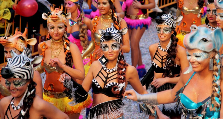 Een kleurrijke carnavalsparade in Sitges, met dansers in extravagante kostuums, veren en glinsterende maskers die door de straten trekken, omringd door feestende toeschouwers.