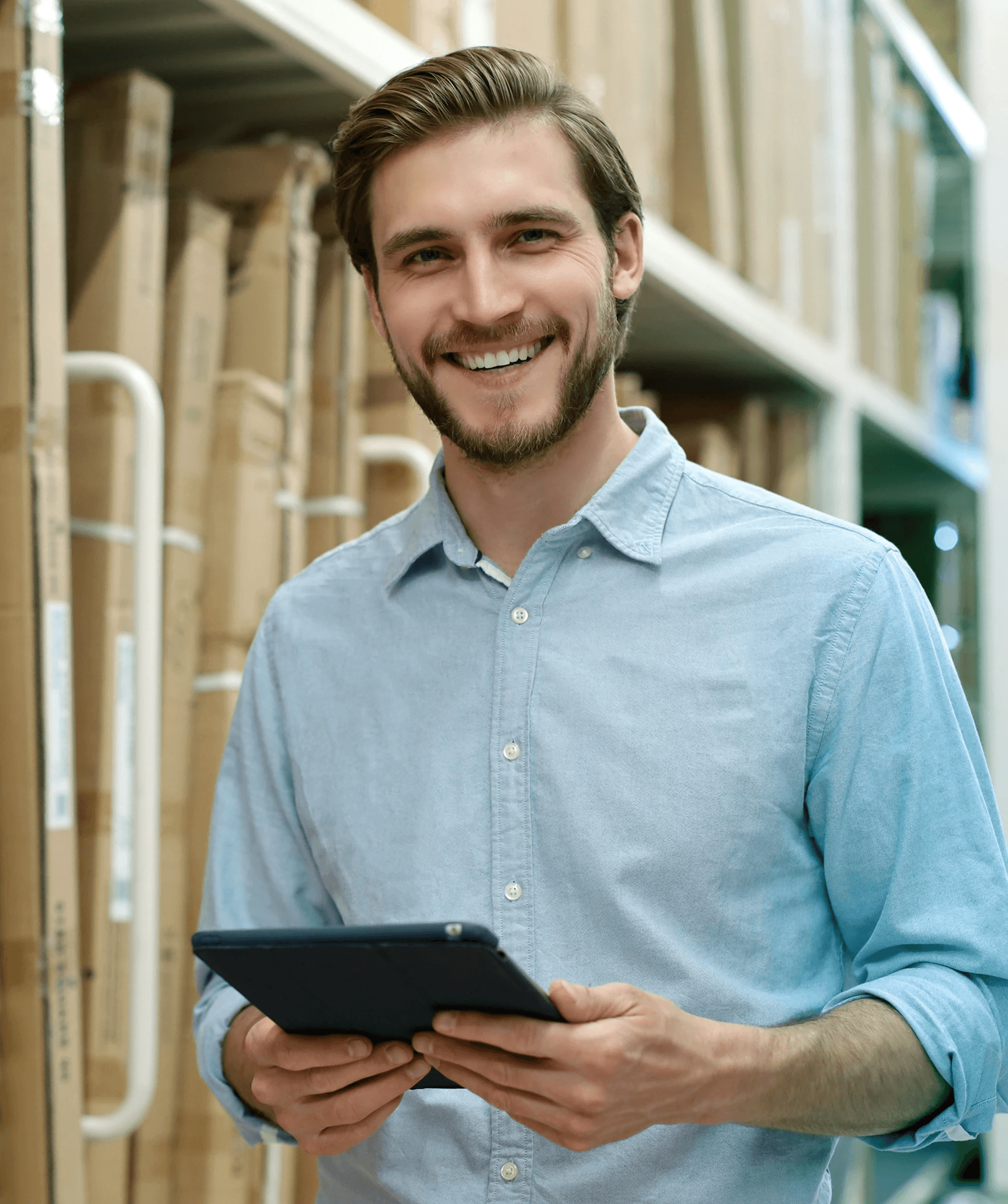 Junger, lächelnder Mann im hellblauen Hemd hält ein Tablet in einem Lager mit Regalen voller Kartons.