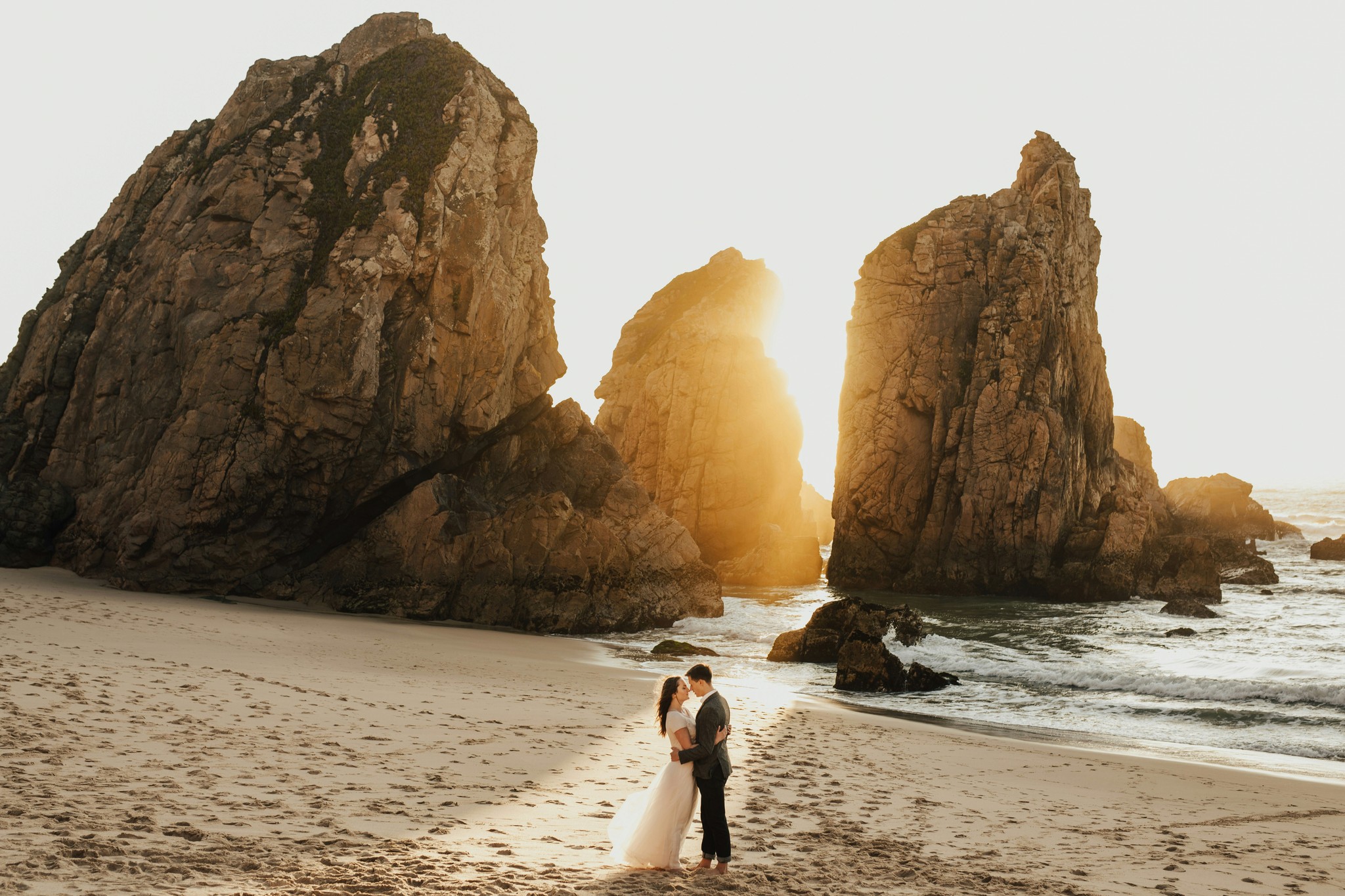 Bride and groom sharing a moment on a coastal beach with towering rock formations silhouetted by the setting sun