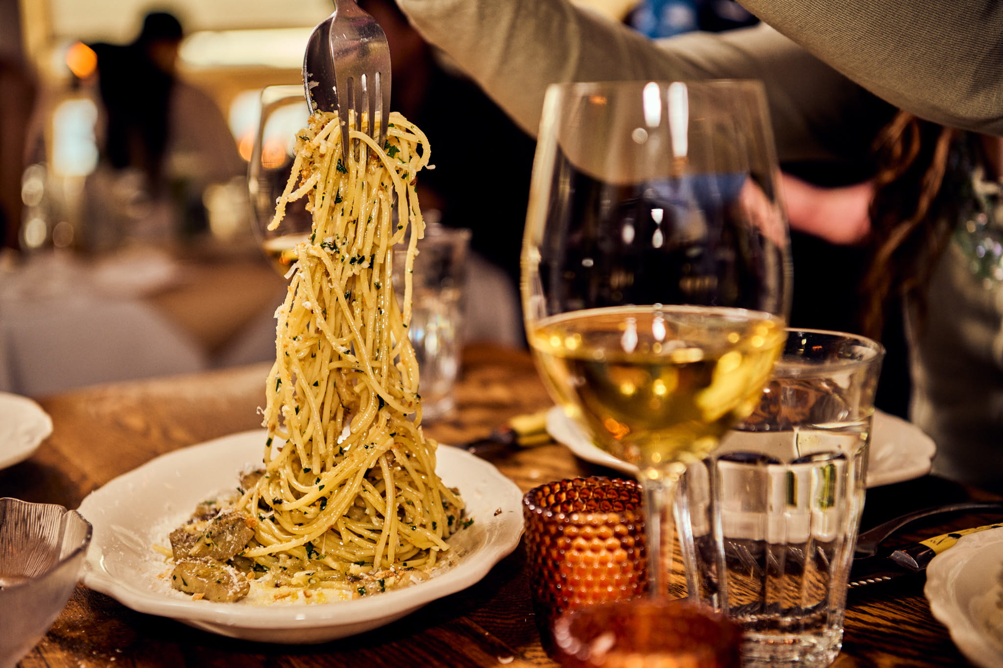 A person lifting a forkful of pasta over a table with a glass of white wine.