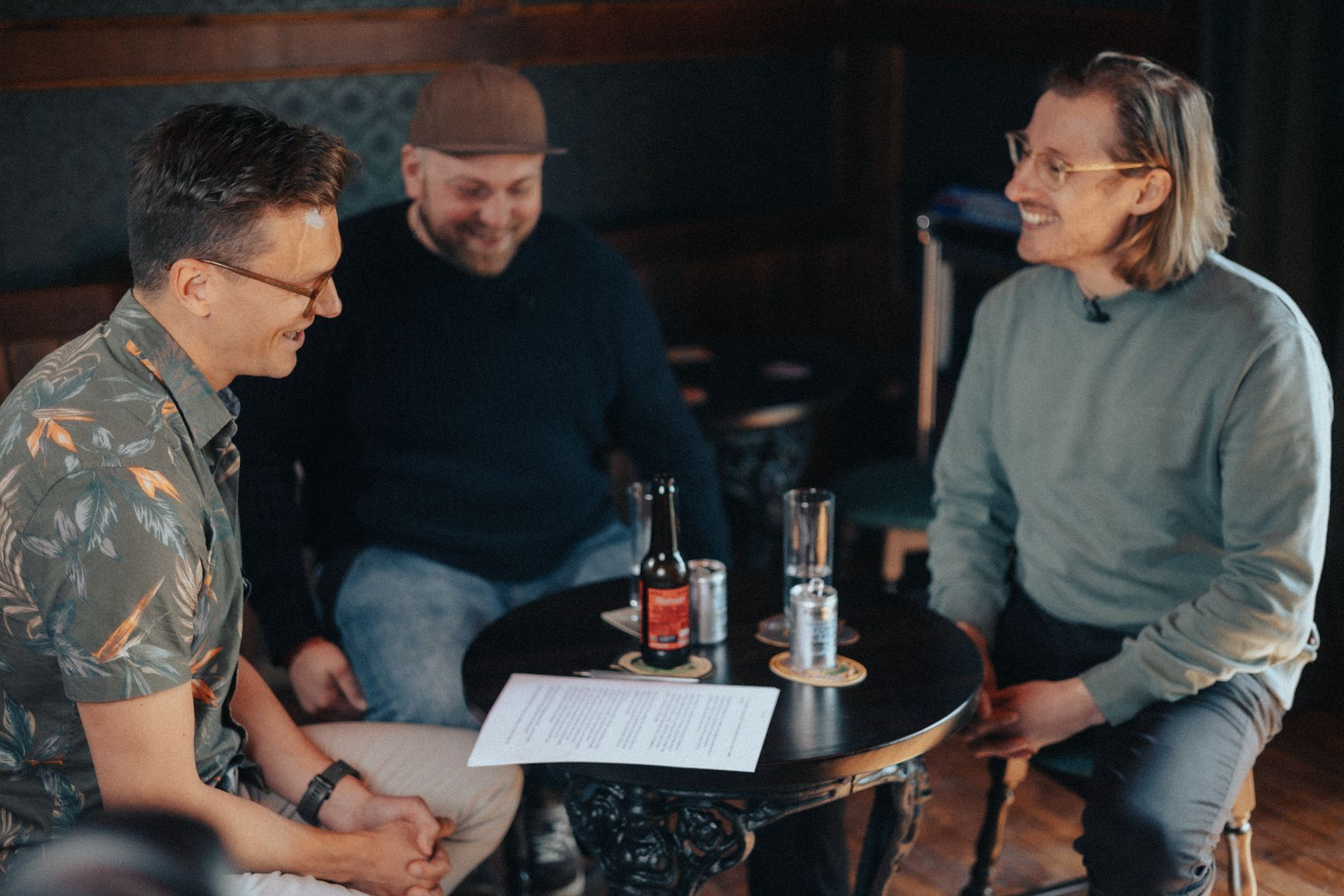 Three men engaged in a discussion, sitting at a table with papers and microphones in a cozy setting.