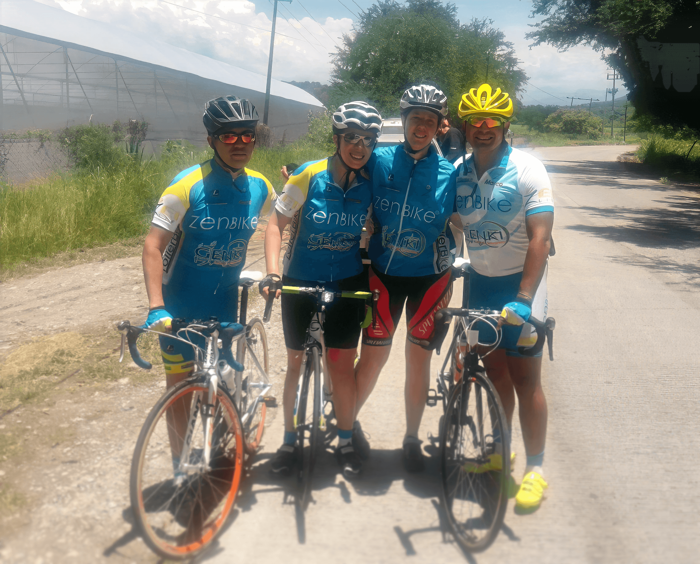 Cuatro ciclistas del equipo zenbike posan en carretera rural con uniforme azul, bajo sol intenso y bicicletas de ruta.