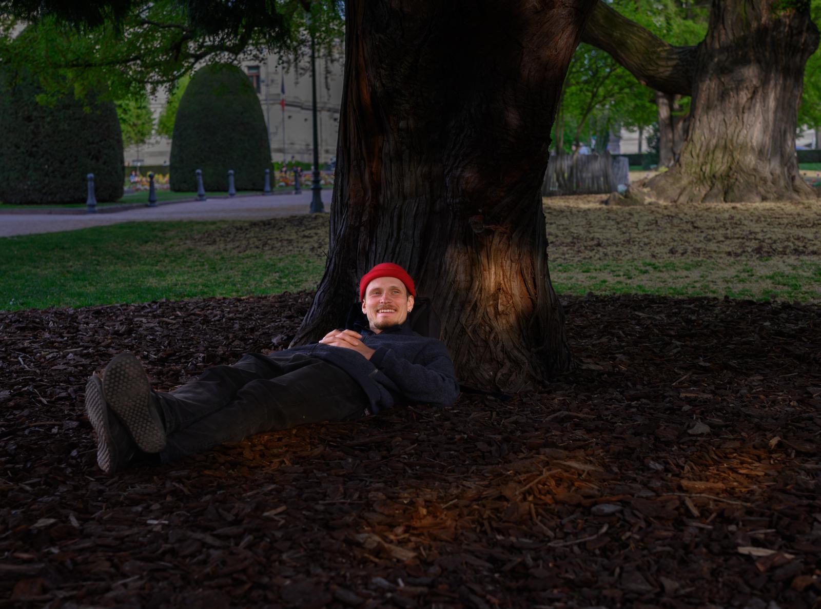 Man liyng under a tree on woodchips  wearing a red hat and a garden with shrubs in the backround.