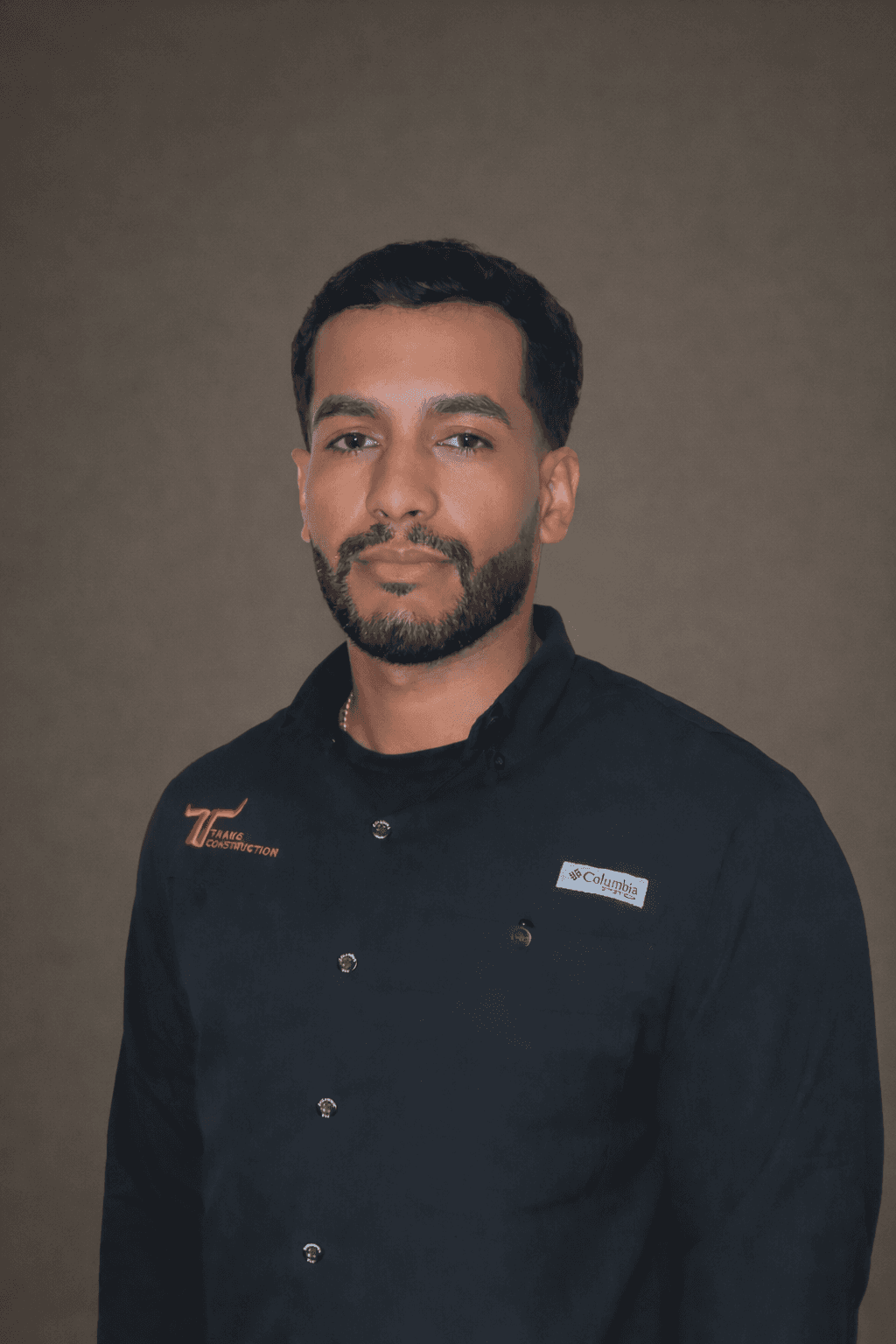 A man wearing a black shirt stands against a neutral background, looking towards the camera with a serious expression.