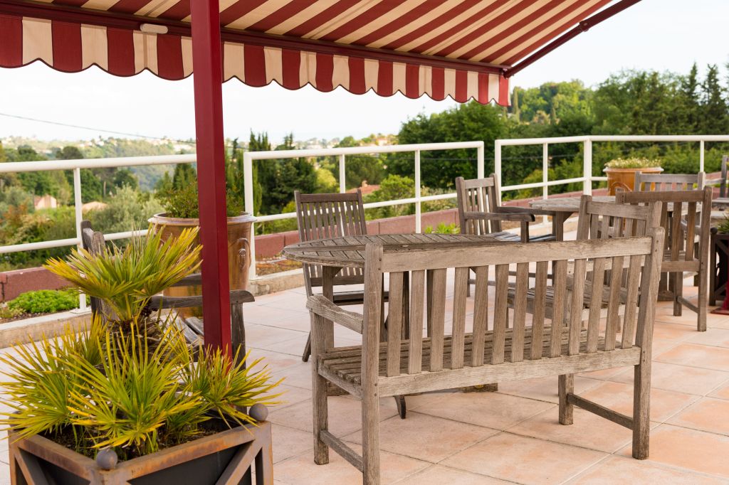 Outdoor terrace with striped red and white awning, wooden seating, tropical plants, and scenic garden views