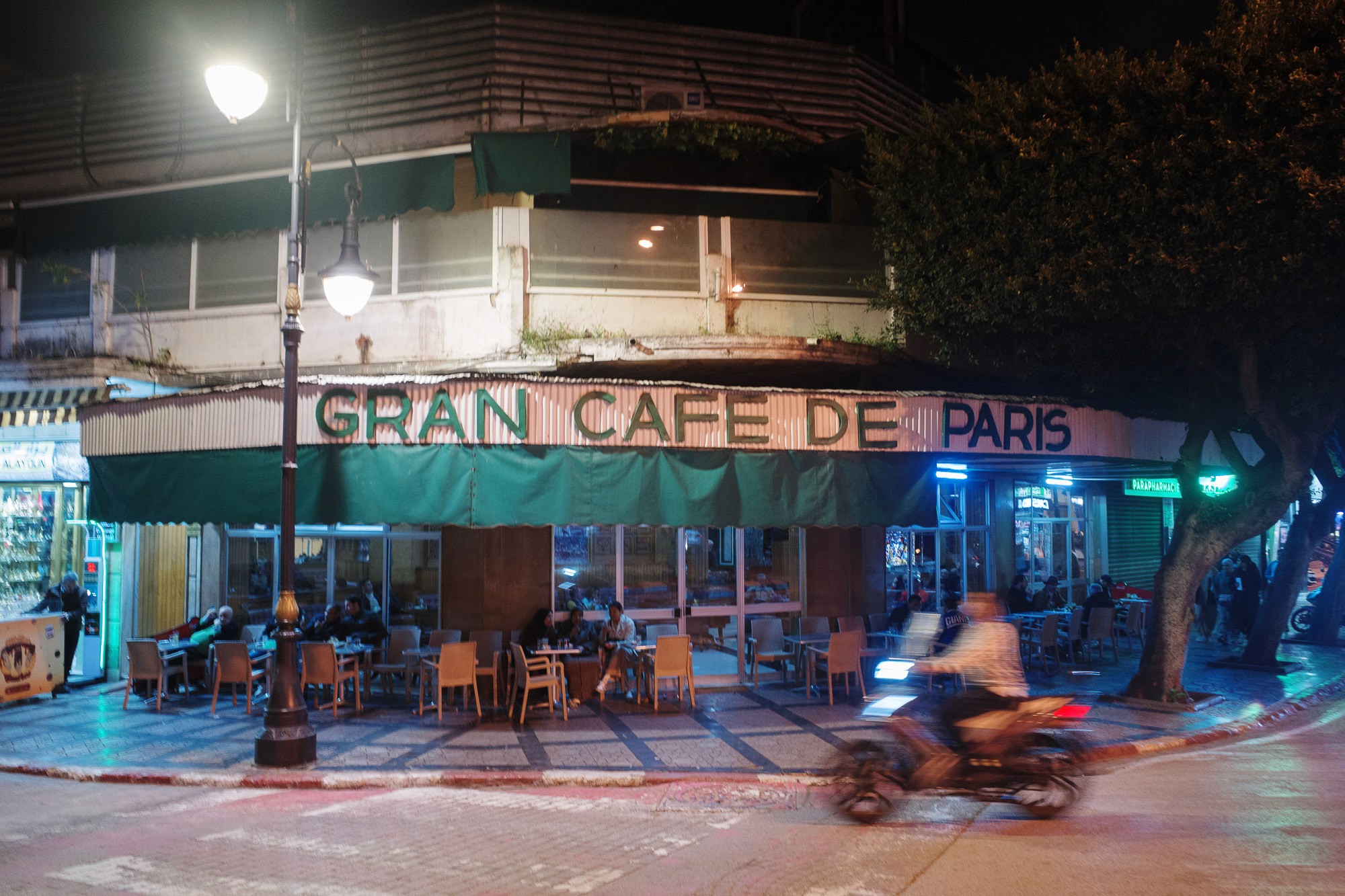A cozy outdoor seating area with patrons dining under a green awning at Gran Cafe de Paris, set against an urban street backdrop with a blurred motorcyclist passing by.