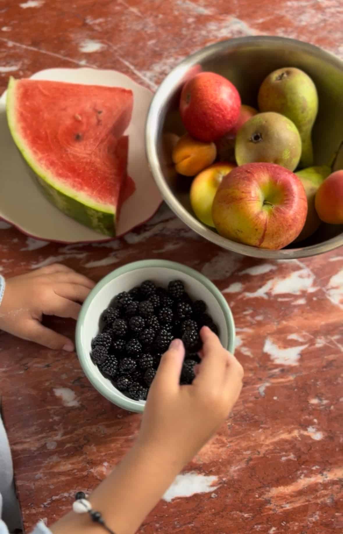 Child’s hands reaching for a bowl of blackberries on a table with watermelon and apples nearby.