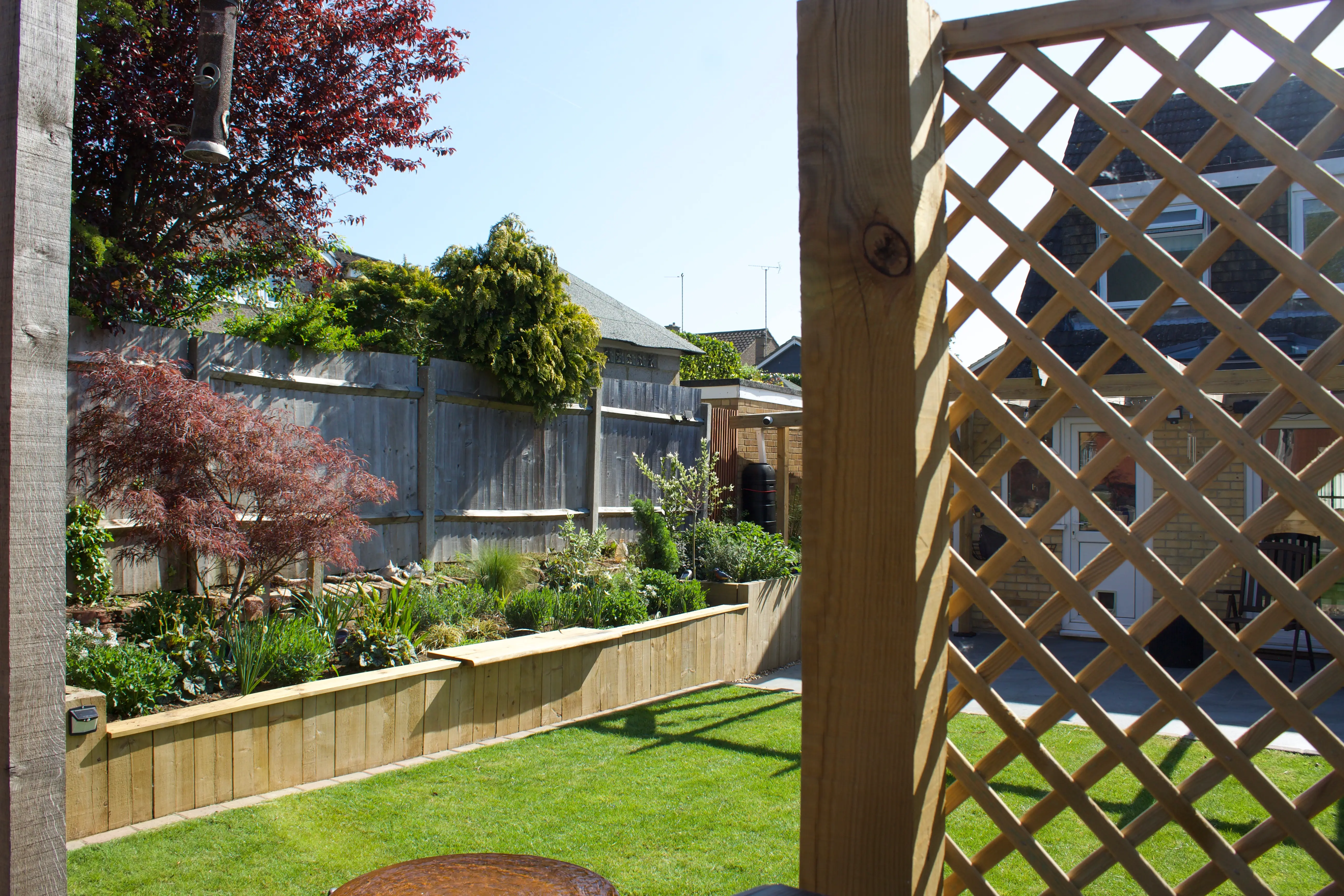 A serene garden view framed by a wooden lattice, featuring greenery and colorful flowers under a clear blue sky.