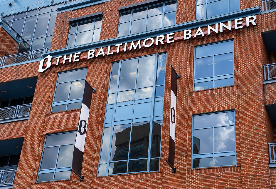 A brick building with large windows displays The Baltimore Banner sign and two banners with a logo.