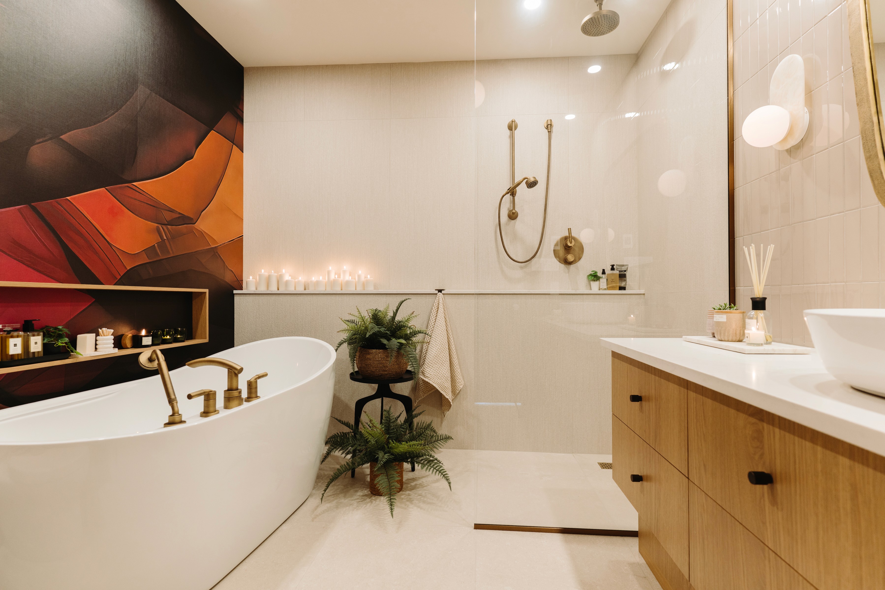 Cette salle de bains moderne et chaleureuse se distingue par sa baignoire autoportante blanche et éléguante, mise en valeur par une murale colorée et une robinetterie dorée. L'espace invite à la détente grâce à ses chandelles allumées, ses plantes verdoyantes et sa douche vitrée épurée. La vanité en chêne naturel se marie très bien parfaitement avec l'ensemble des teintes chaudes présentes dans la pièce.