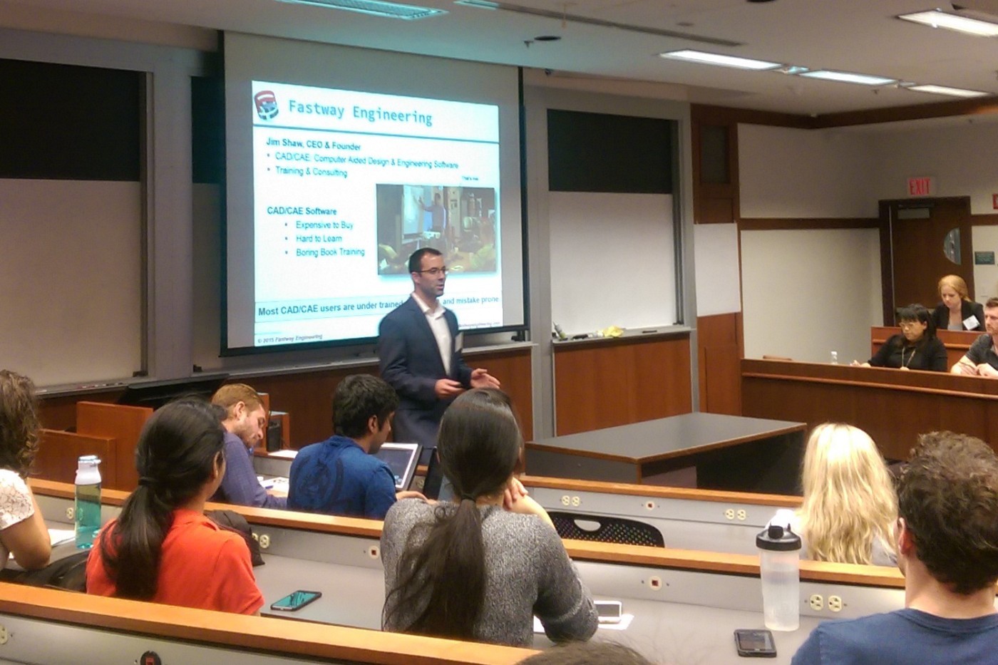 A group of people sitting in a classroom with a man standing in front, presenting on a projector screen. The presentation highlights CADCAE software and its applications, including engineering design, training, and workflows, alongside a small group of students.