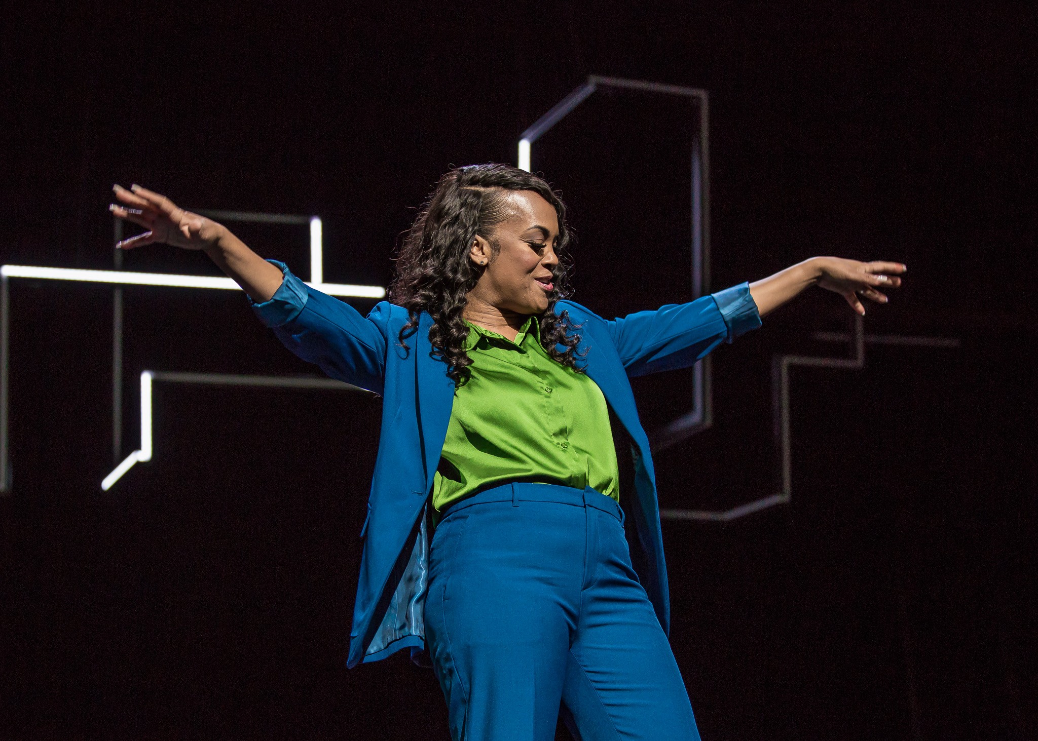 A person wearing a vibrant outfit strikes a dynamic pose on stage, with colorful lighting in the background.