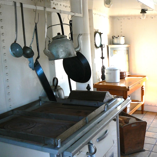 A vintage kitchen with hanging utensils, a teapot, a stove, a wooden table, and various kitchenware on surfaces and shelves.