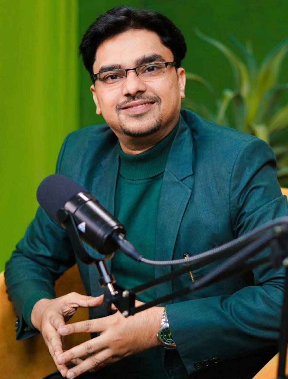 A young man with glasses and a denim shirt sits confidently against a blue wall. Art and cacti decorate the space, conveying a creative vibe.
