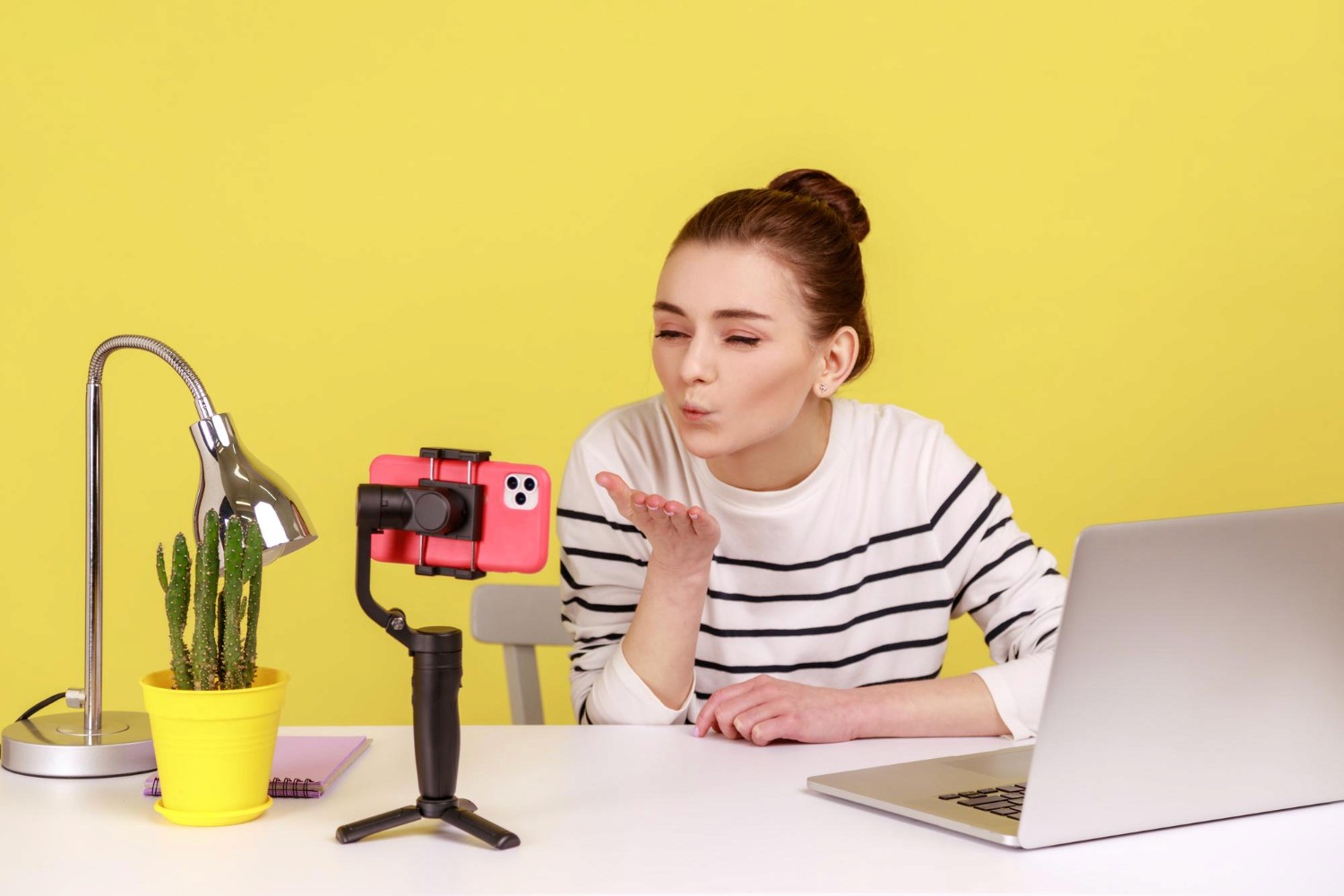 A woman seated at a desk, working on a laptop with a camera positioned nearby, focused on her tasks.