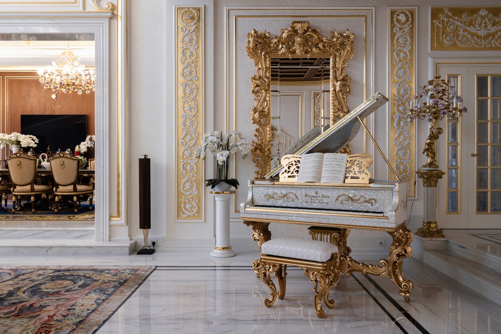 A luxurious, ornate room features a lavish white and gold grand piano with intricate carvings, set on polished marble flooring, surrounded by elegant decor including a large golden mirror and an opulent chandelier in the adjacent dining area.