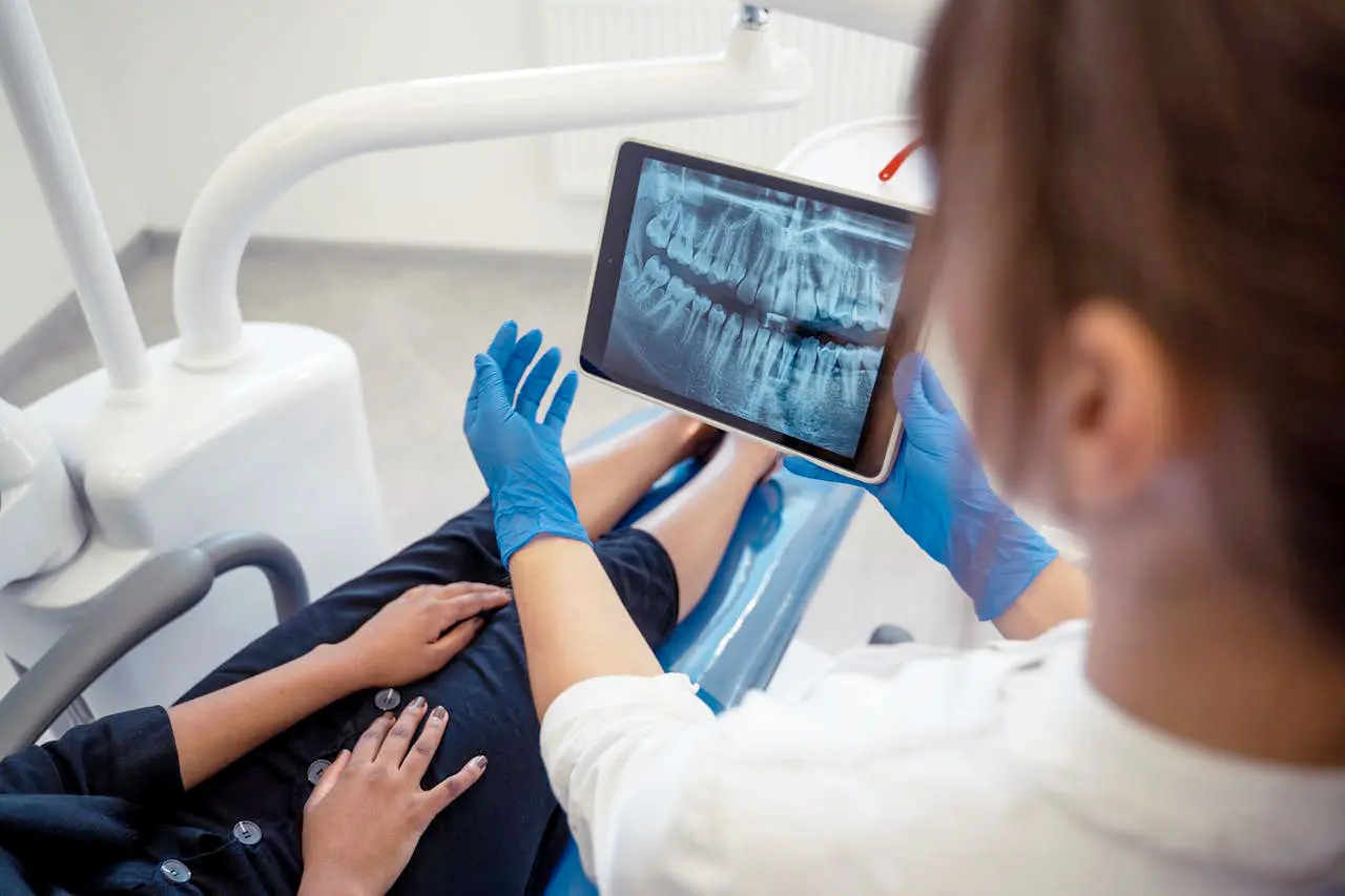 A dental professional shows a patient an X-ray on a tablet while seated in a dental office.