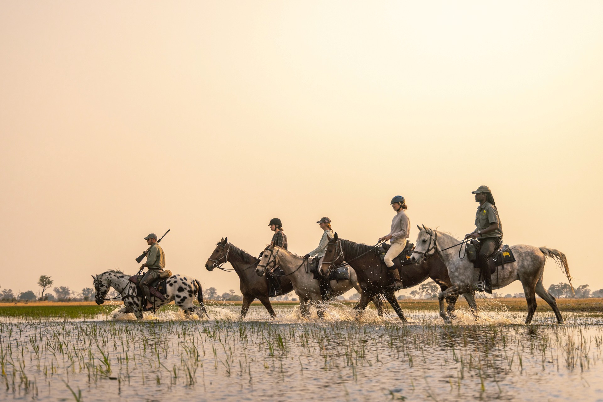 Kujwana Delta Ride, Okavango Delta Botswana. Fem ryttare till häst på ridsemester vadar genom grunt vatten i skymning.