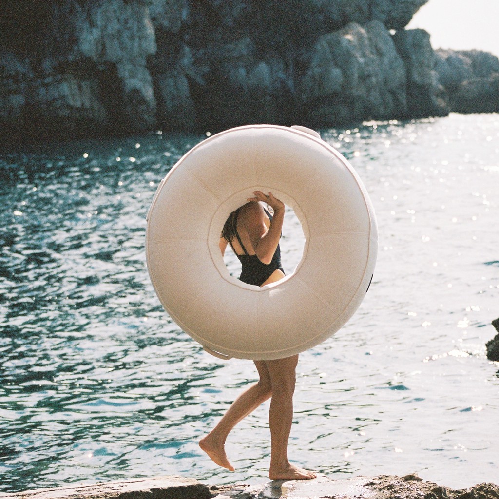 Woman carrying a luxury white pool float along a sunlit Mediterranean coastline with sparkling sea