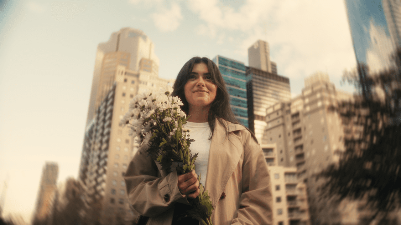A woman holding a bouquet of flowers, smiling and standing in a park with tall city buildings in the background