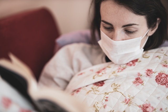 Photo of woman with respirator mask sitting in bed reading a book