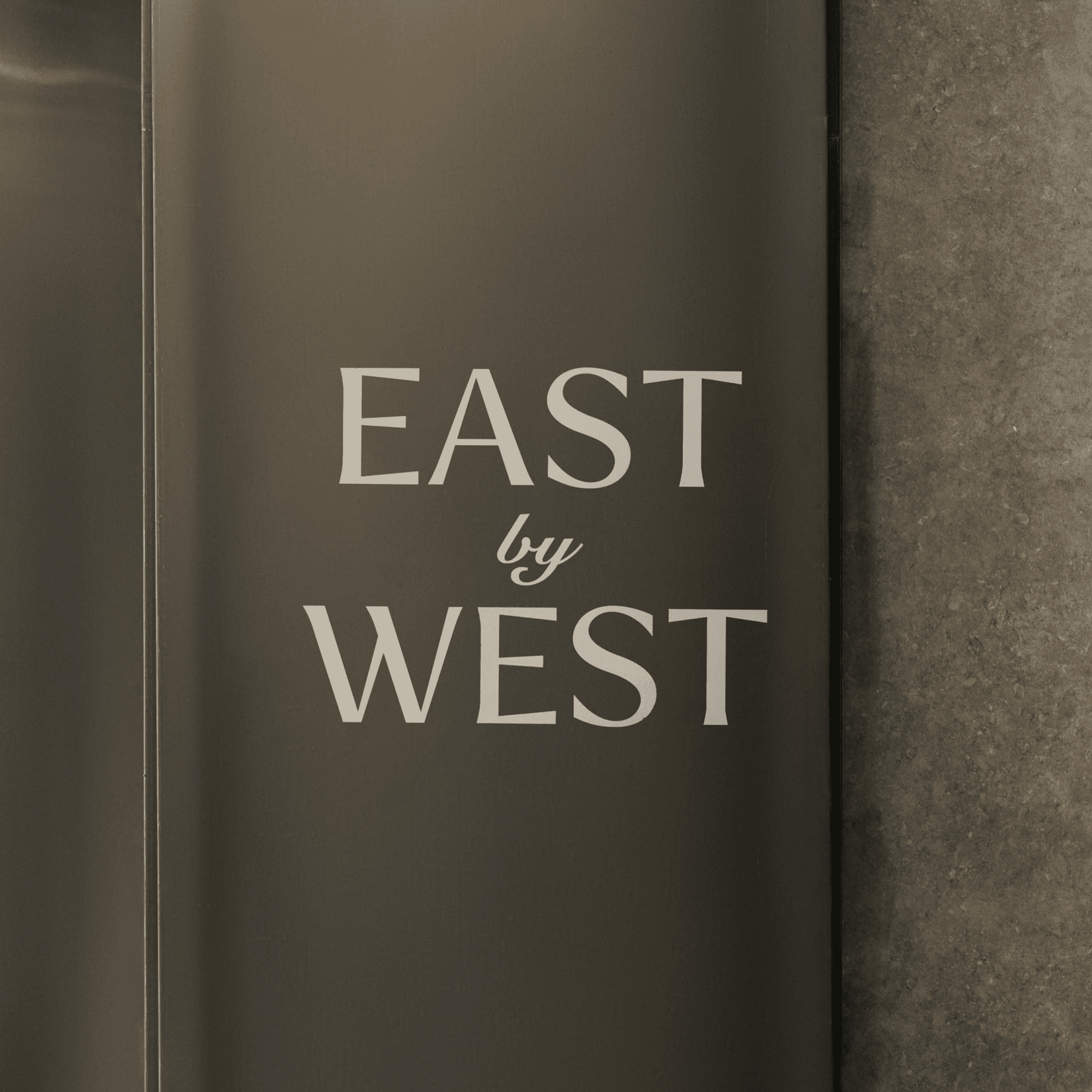 East by West restaurant signage in elegant sans-serif font on sleek dark gray wall flanked by warm-toned vertical wood slats and metallic panel. 